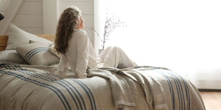 Woman sitting on bed with Swans Island grey blanket with blue stripes, daydreaming with a bright wind behind.