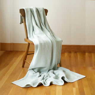 A pale mint green textured knit throw blanket draped over a wooden chair in a room with light wood floors and a cream wall.