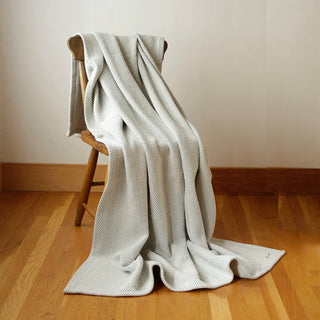 A light gray textured throw blanket with a fine herringbone pattern draped over a wooden chair in a room with light beige walls and a wooden floor.