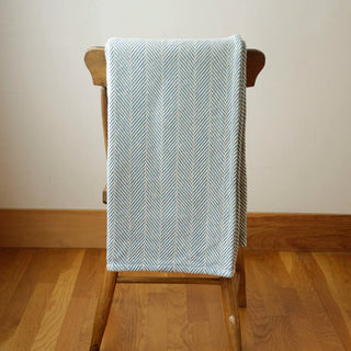 A light blue and white herringbone patterned throw blanket draped over a wooden chair in a minimalist room with a beige wall and wooden floor.