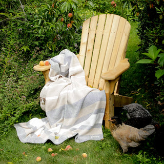 A light natural wood Adirondack chair in a garden setting with a Sand and Sea striped cream, beige, and gray cotton throw blanket draped over it, surrounded by green foliage, fallen fruit, and two chickens.