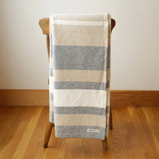 A striped woven throw blanket draped over a wooden chair in a room with light wood floors and a plain wall. The blanket features horizontal stripes in cream, beige, and slate blue-gray, with a small white woven Swans Island brand label near the bottom.