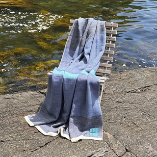 A blue handwoven blanket with a turquoise stripe and cream trim is draped over a wooden chair beside a rocky shoreline with water in the background.