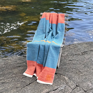 A handwoven throw blanket with color-blocked blue, coral, and cream sections draped over a wooden beach chair next to a body of water.
