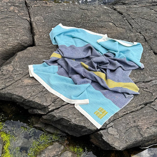 A draped striped handwoven throw blanket  in light aqua, dark marine blue, and fern green, with white silk trim and a green handwoven logo, laid out on dark granite rocks outdoors by the harbor.