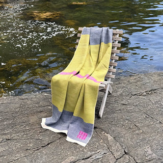 A handwoven throw blanket in fern green marine blue, and fuchsia pink is draped over a wooden beach chair outdoors next to the Maine ocean.