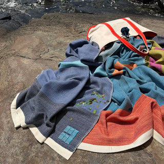 A pile of colorful handwoven throw blankets and a cream tote bag with red handles resting on a rocky outdoor surface near water.