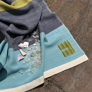 A close-up of a handwoven throw, laid on a rough stone surface. The fabric has a color-blocked design with dark marine blue at the top, light aqua blue at the bottom, and a cream border. 