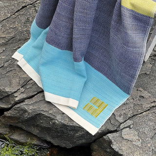 A closeup detail of a handwoven Swans Island throw blanket with a color-blocked pattern of navy blue, light blue, and white, featuring a fern green woven logo, draped over dark gray rocks outdoors by a Maine harbor.