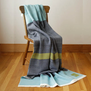 A striped handwoven blanket in light blue, dark blue, and fern green is draped over a wooden chair on a hardwood floor against a plain wall.