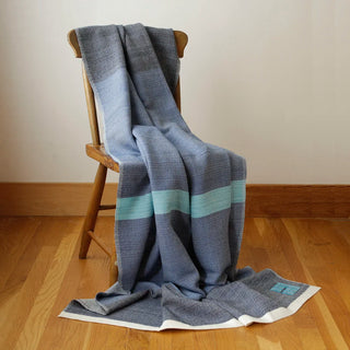 A woven throw blanket in shades of dark marine blue, light blue, aqua, and white, draped over a wooden chair on a hardwood floor against a neutral wall.