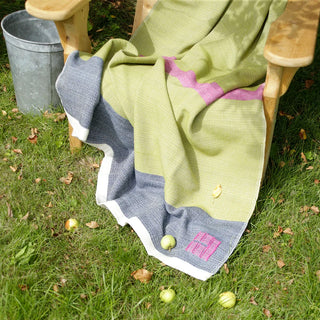 A large handwoven Swans Island throw blanket with a color-blocked design in fern green, dark marine blue, and fuchsia pink, draped over a wooden outdoor chair on a grassy lawn with scattered green apples.