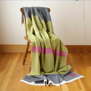 A striped handwoven blanket in fern green, dark marine blue and fuchsia pink is draped over a wooden chair in a room with light wood floors and a cream wall.