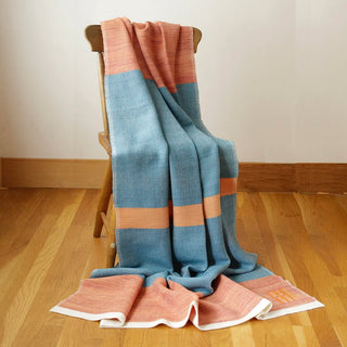 A woven throw blanket with wide stripes of azure blue, terracotta orange, nasturtium orange with a white border, draped over a wooden chair on a light wood floor against a neutral wall.