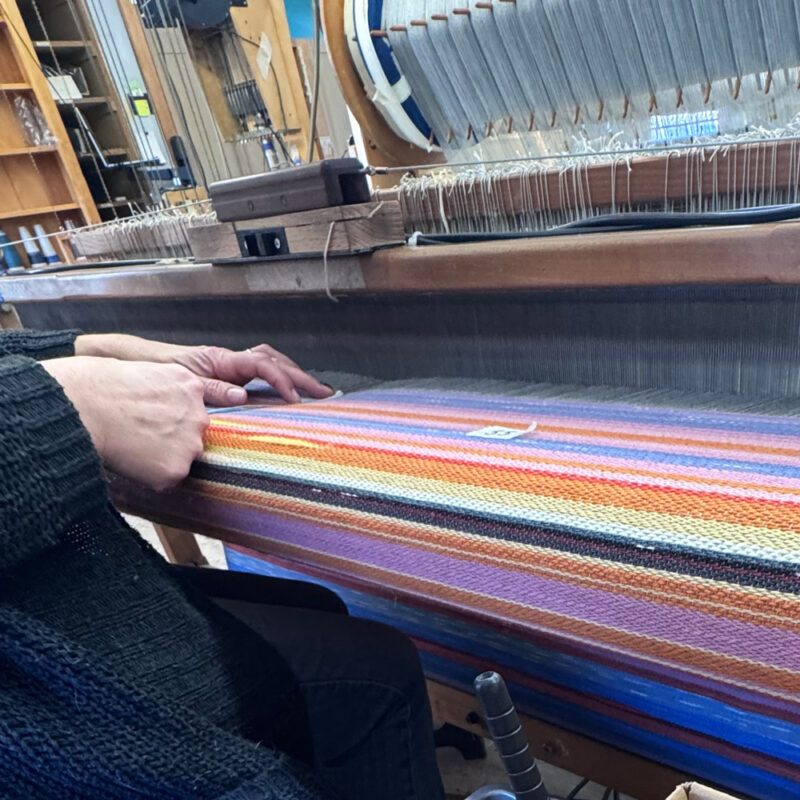 Swans Island Company's Horizon Limited Edition 2026 Throw - A tangible homage to the vibrant colors of first light and golden hour when the sun rises and sets. Handwoven in Maine with hand-dyed wools - a textiles collectors piece. A Swans Island artisan weaver at the loom making a Horizon Limited Edition throw.